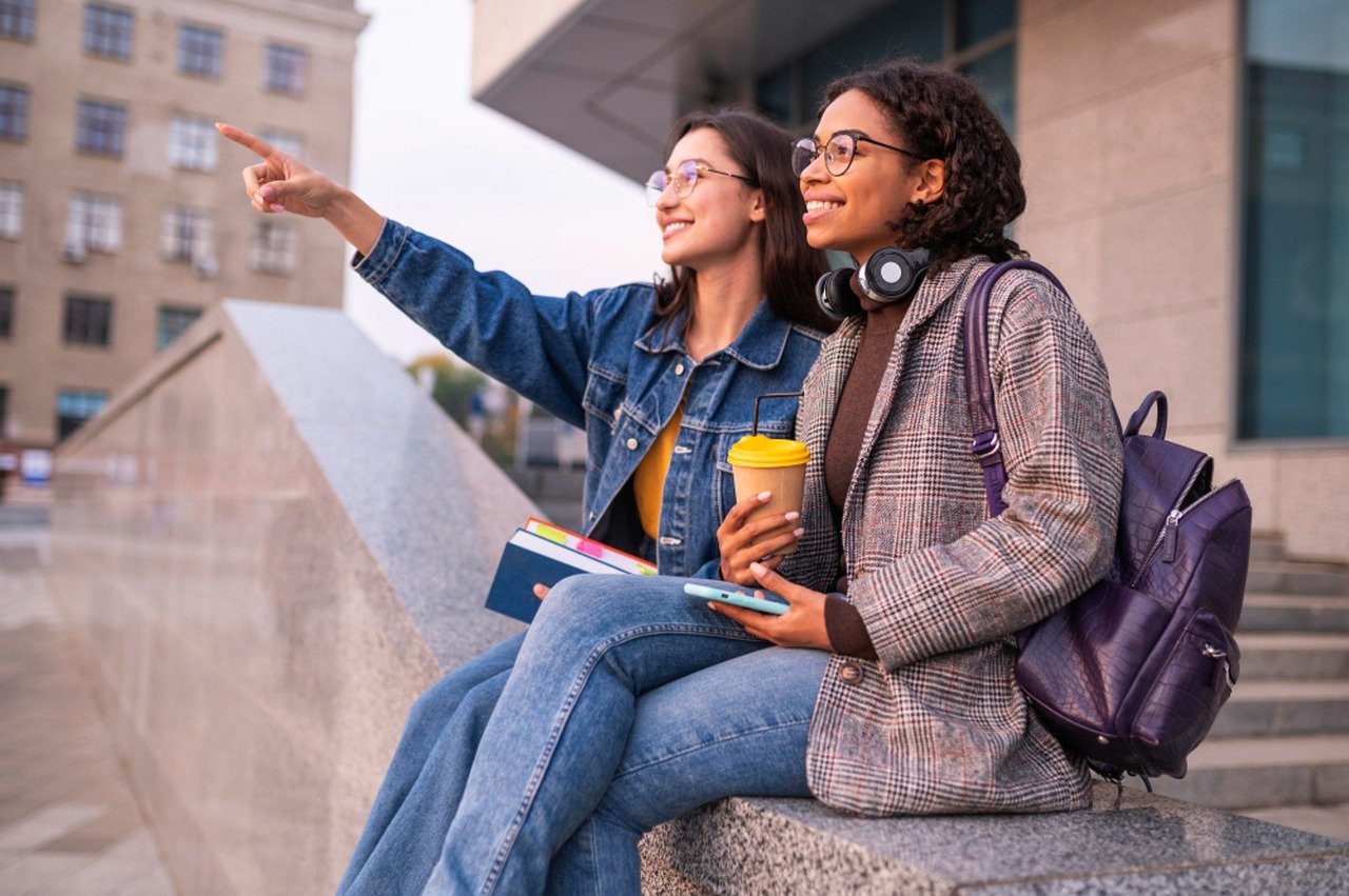 Photo de deux étudiantes à Paris, assises à l'extérieur d'un campus parisien, près de leur logement étudiant Paris 20. L'une d'elles montre du doigt quelque chose, elles sont toutes les deux heureuses.