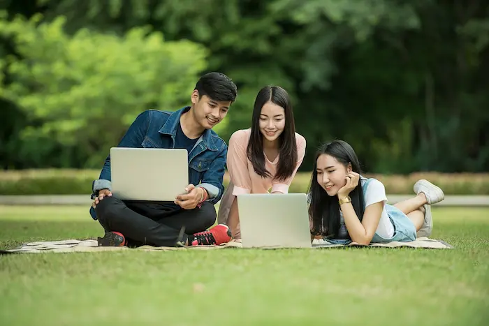 Photo de deux étudiantes et d'un étudiant assis dans l'herbe avec leurs ordinateurs. Ils travaillent dans l'espace extérieur de leur université, près d'un logement étudiant Toulon