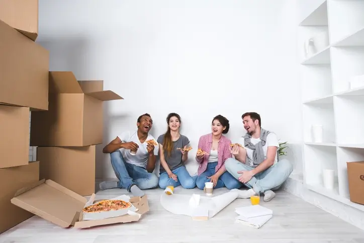 Des étudiants assis au milieu de cartons et mangent une pizza, contents d'emménager dans leur nouveau logement en colocation à Evry