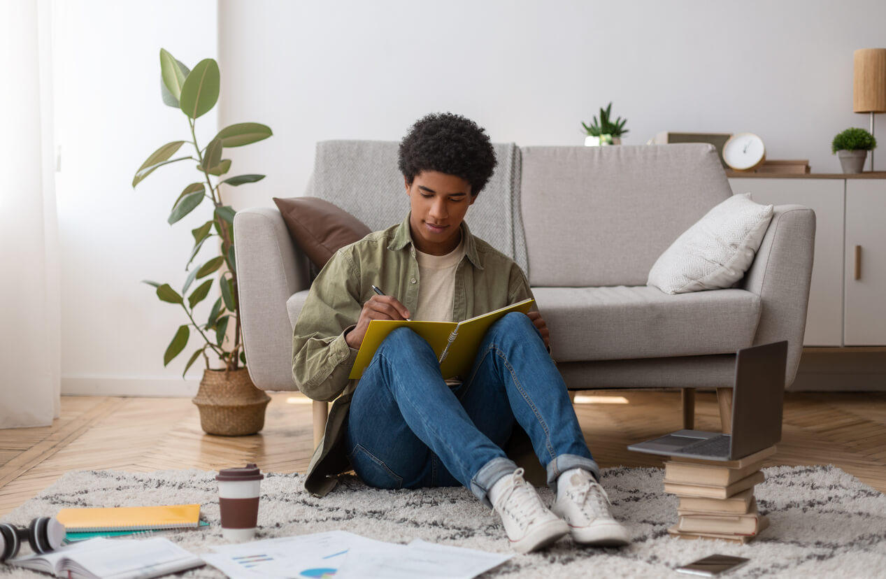 Photo d'un étudiant assis sur le sol de son salon, dans une résidence étudiante à Paris 18. Il écrit sur un carnet, des livres et des cahiers sont éparpillés autour de lui sur le tapis.