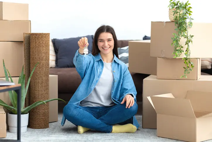 Photo d'une étudiante assise dans le salon de son nouveau logement en location étudiant. Elle sourie et regarde la caméra, montrant la clé de son logement étudiant en location.